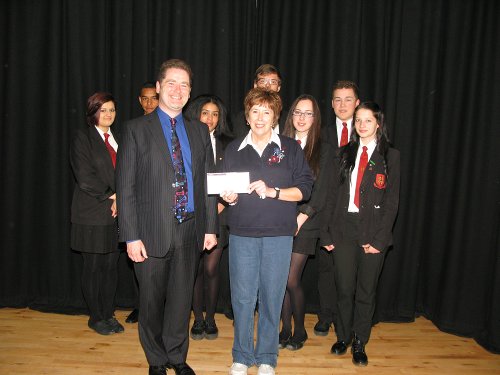 Headmaster Mr presents Miss Allan with the cheque whilst a group of year 11 pupils look on.